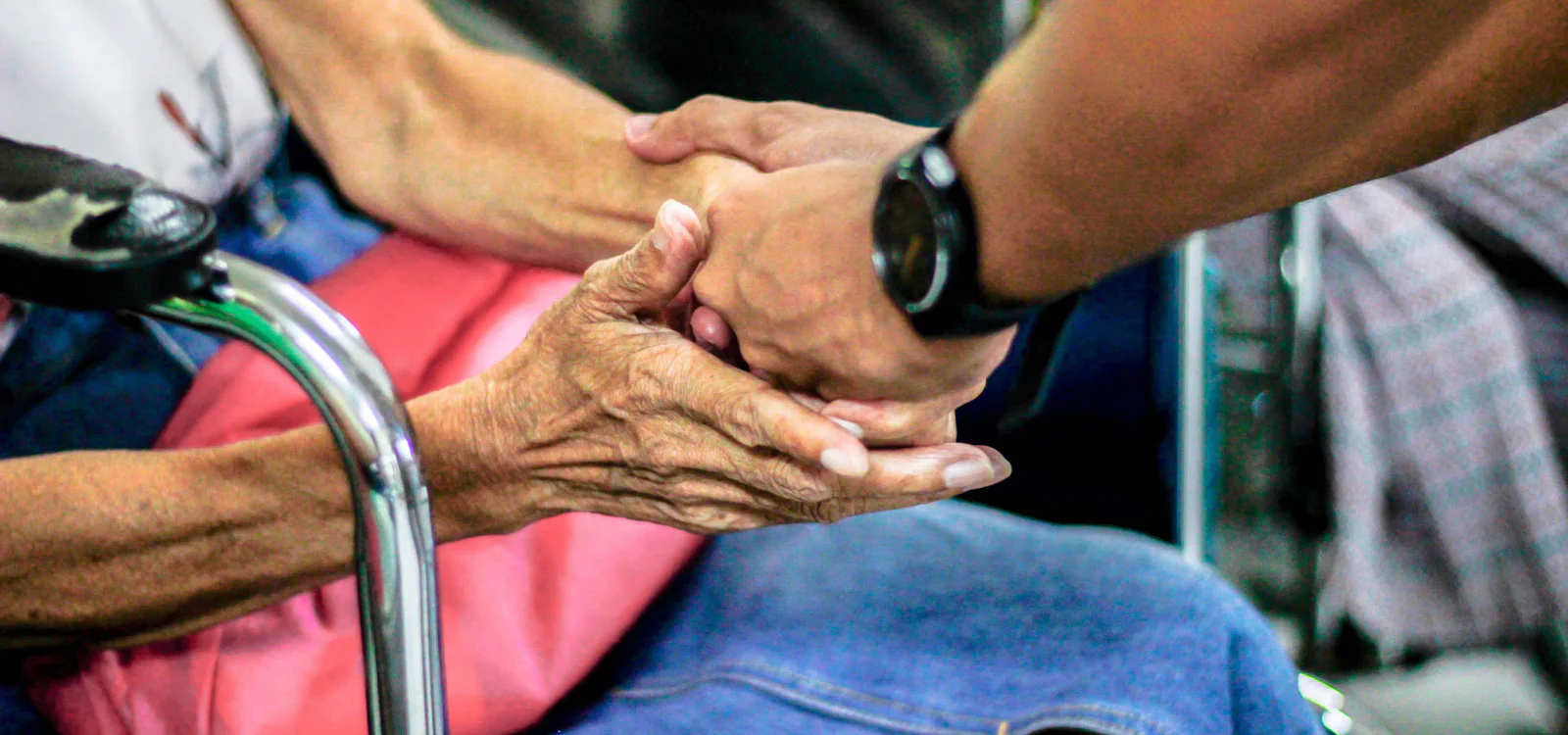 A younger hand holding an older person's hand in both of theirs, beside a walking frame